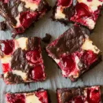 Close-up of cherry cheesecake brownies cut into squares, showing swirls of cream cheese and whole cherries in a rich chocolate base.