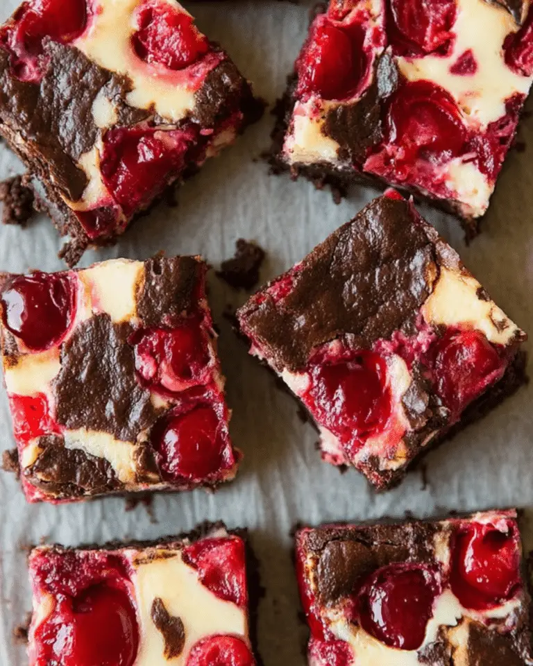 Close-up of cherry cheesecake brownies cut into squares, showing swirls of cream cheese and whole cherries in a rich chocolate base.