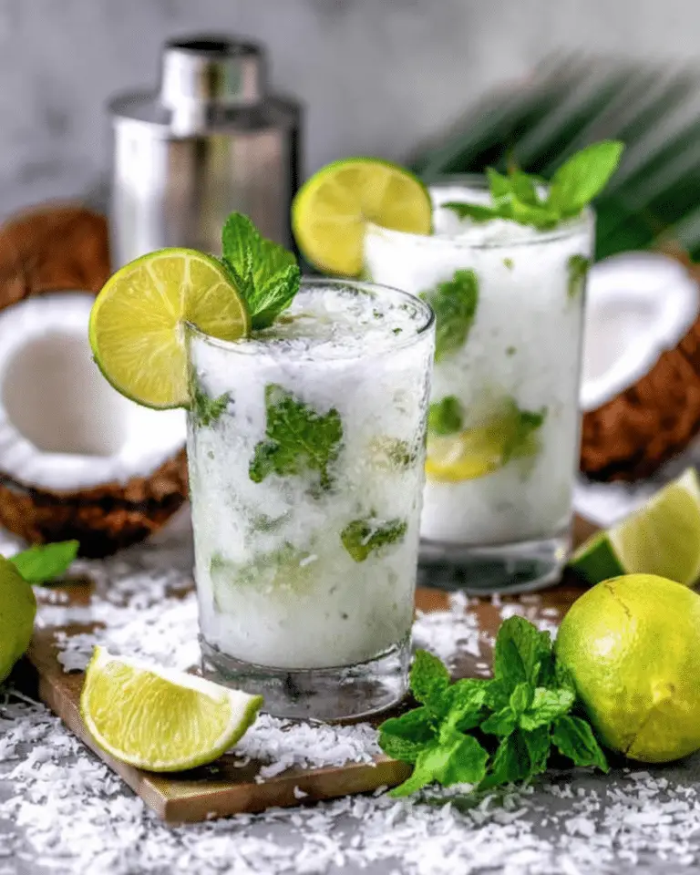 Two glasses of coconut rum mojitos garnished with lime slices and mint, surrounded by fresh limes, mint, and shredded coconut.