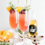 Two cranberry orange mimosas garnished with fresh cranberries, orange slices, and rosemary sprigs in champagne flutes.