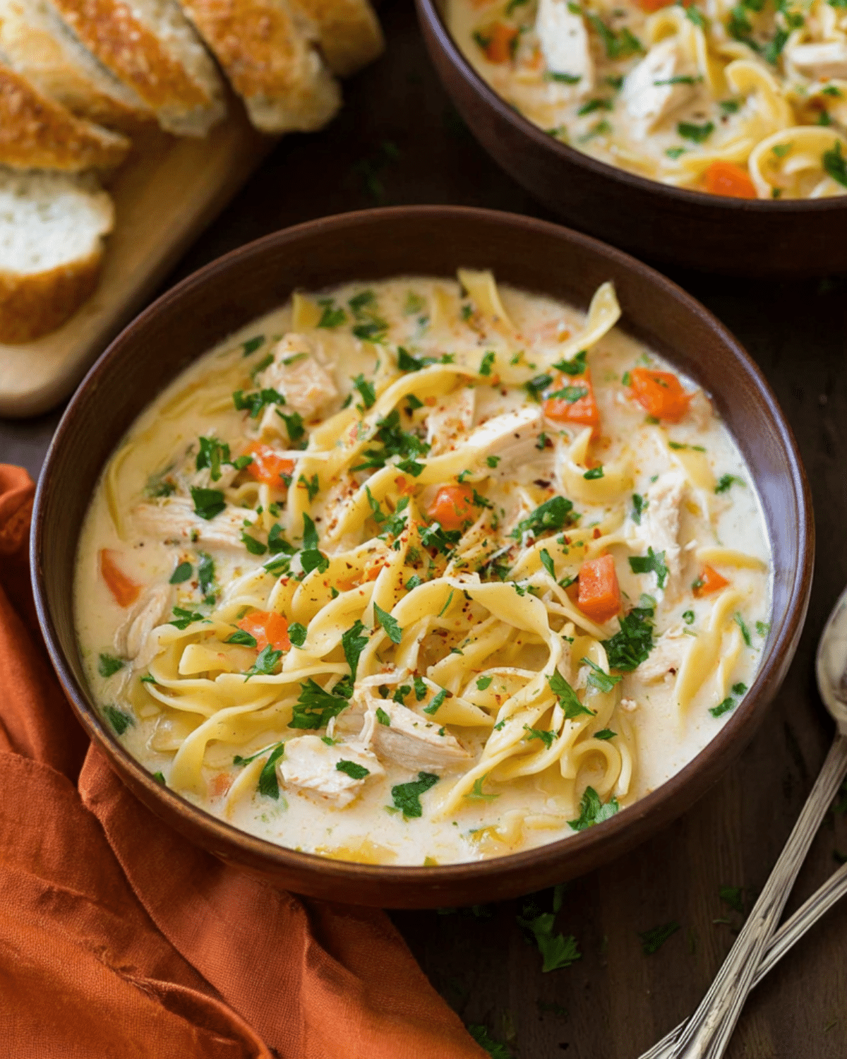 Bowl of creamy chicken noodle soup with egg noodles, shredded chicken, carrots, and herbs, served with sliced bread.
