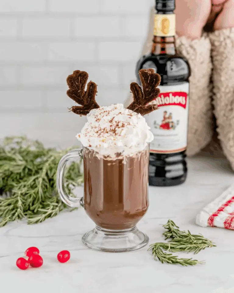 Glass mug of hot chocolate topped with whipped cream, chocolate antlers, and cinnamon, labeled "Drunken Rudolph," with a bottle of liqueur and festive decor in the background.