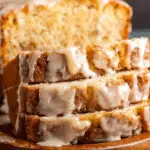 Slices of eggnog bread drizzled with glaze, stacked in front of a loaf on a wooden serving board.