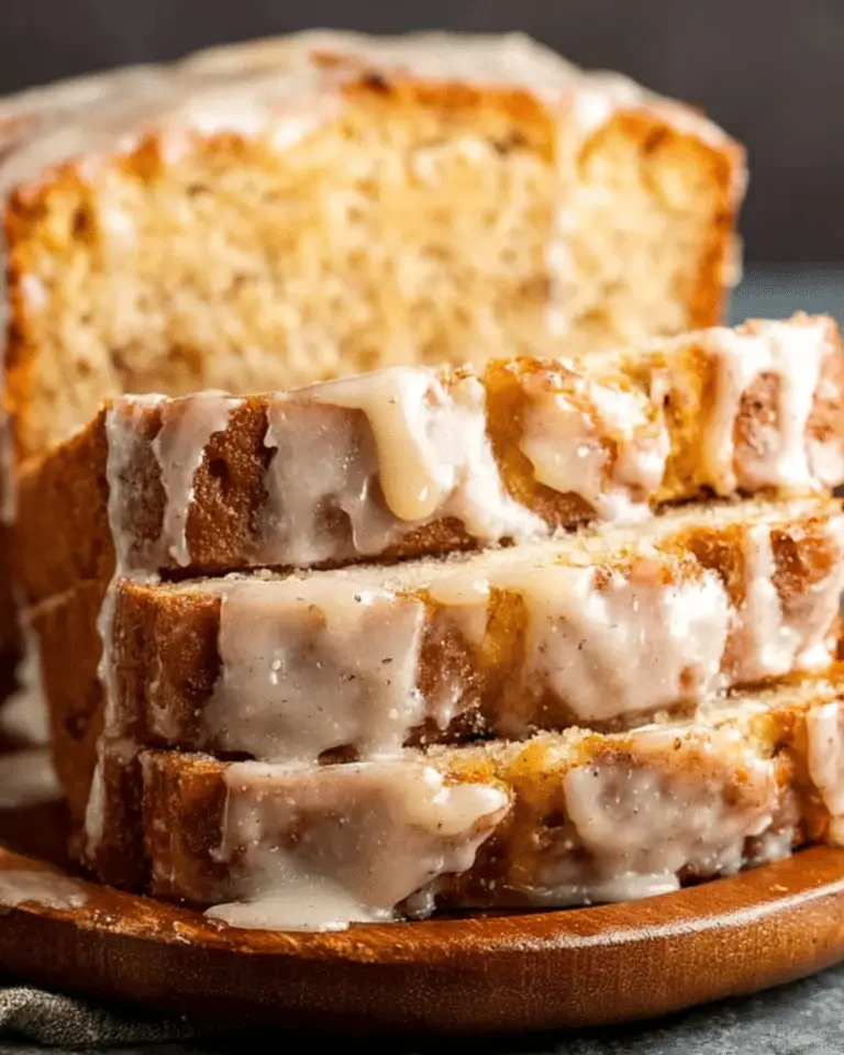 Slices of eggnog bread drizzled with glaze, stacked in front of a loaf on a wooden serving board.