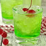 A vibrant green Grinch cocktail served over crushed ice in a glass, garnished with two red maraschino cherries.