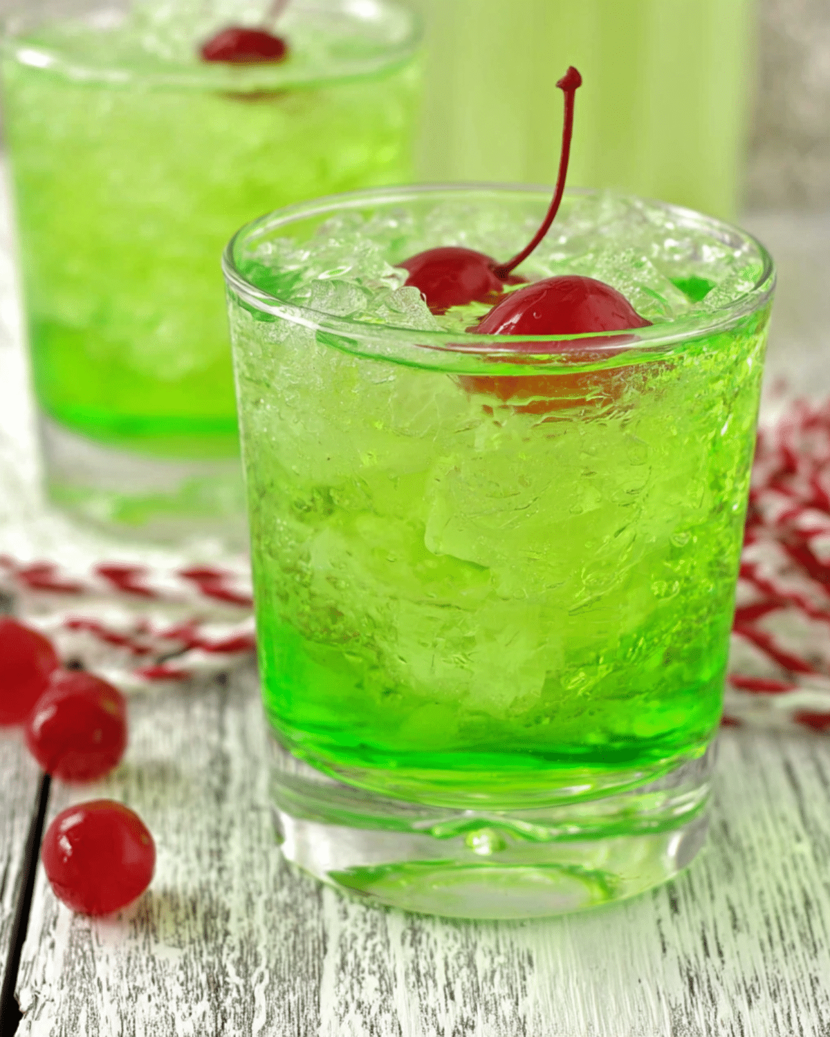 A vibrant green Grinch cocktail served over crushed ice in a glass, garnished with two red maraschino cherries.