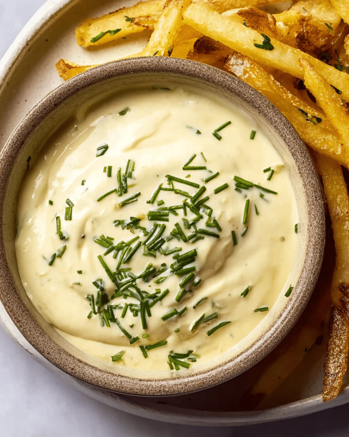 Bowl of homemade garlic aioli topped with chopped chives, served alongside crispy French fries.