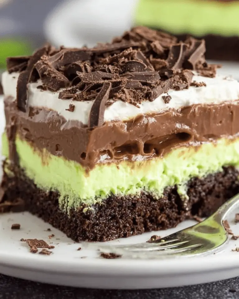 A slice of mint chocolate lasagna dessert with layers of chocolate crust, mint cream, chocolate pudding, whipped topping, and chocolate shavings.