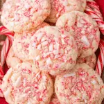 A pile of peppermint cookies topped with crushed candy canes on a red cloth, surrounded by whole candy canes.